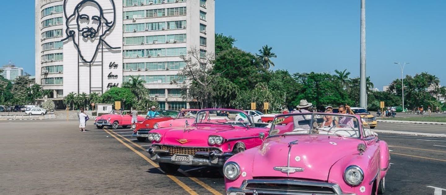 Pink convertibles parked in front of building decorated with iron outline of Camilo Cienfuegos’ face